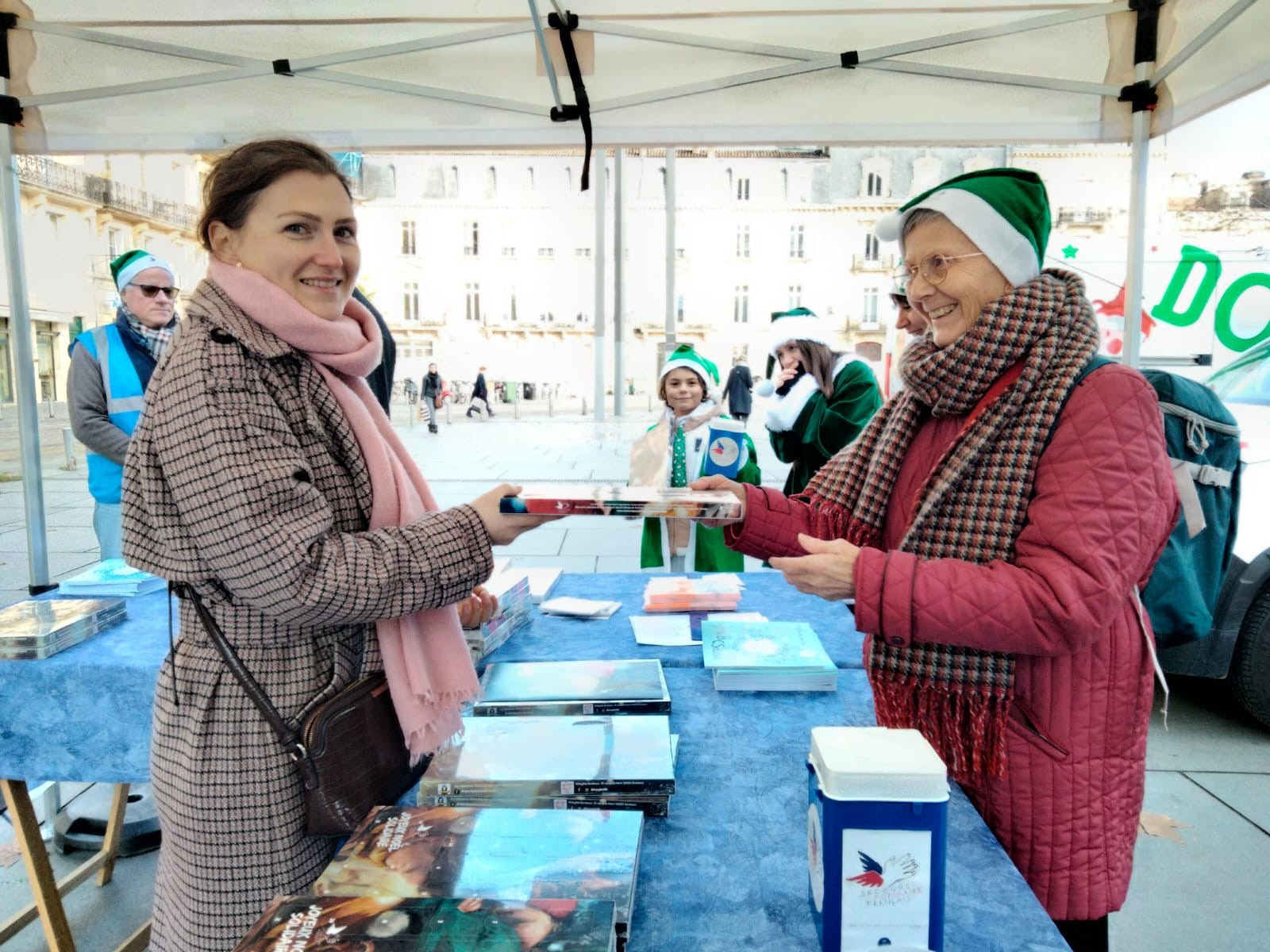 © Cassandre Thomas. Annie Garat (à droite) vend un calendrier, lors du passage de la caravane du Père Noël. Place Pey Berlan, Bordeaux, le 6 décembre 2023. Annie Garat présente "Copains du monde"