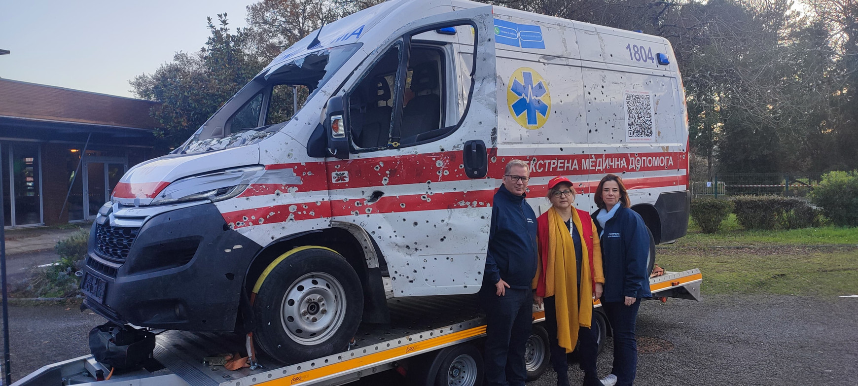 © Cassandre Thomas. Svitlana Poix aux côté de deux pompiers solidaires devant l'ambulance mitraillée, de passage à Bordeaux du 30 novembre au 1er décembre 2024. Ambulance mitraillée : la violence de la guerre en Ukraine exposée à Bordeaux